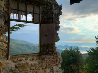 View of lush green mountains and trees through the ruins of a stone wall with a broken window frame on Troodos Mountain.